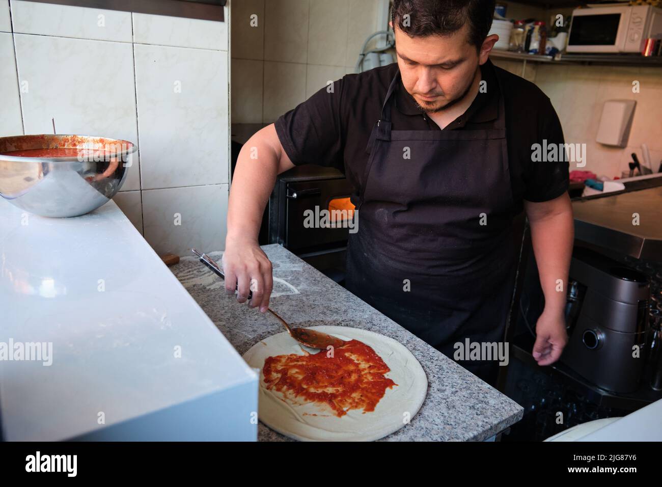 Lateinischer Mann, der in einer Restaurantküche Tomatensauce zu einem Pizzateig hinzufügt. Stockfoto