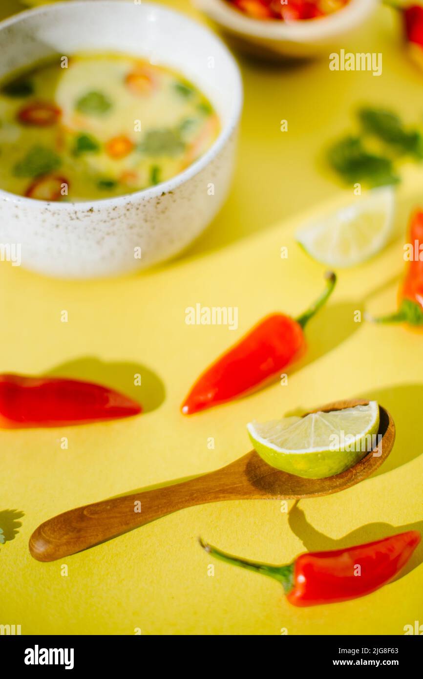 Ein selektiver Schuss Zitrone, Chilis auf gelbem Tisch mit einer Schüssel thailändischer Kokosnusssuppe im Hintergrund Stockfoto