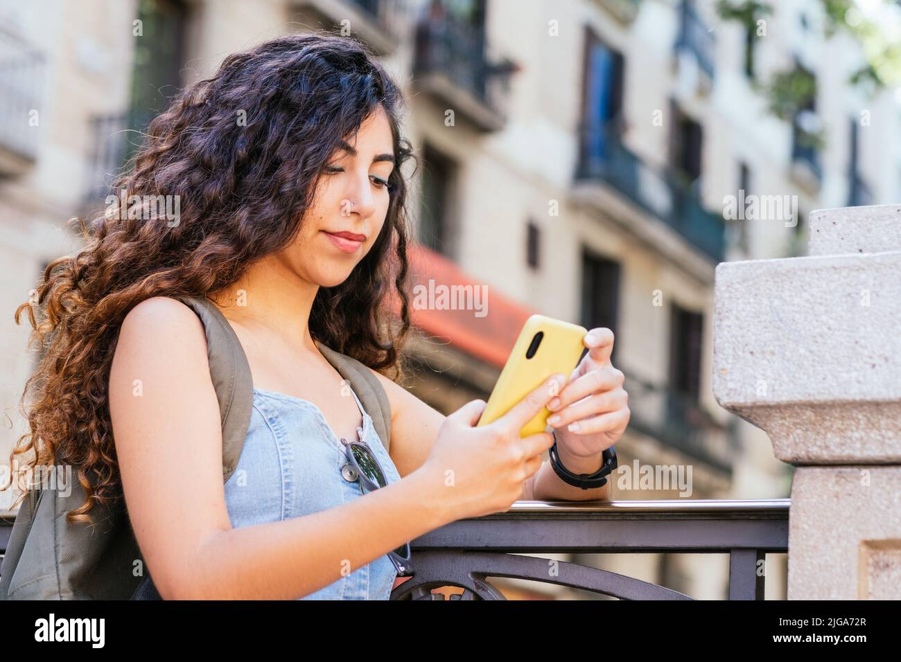 Junge türkische Touristin mit einem Rucksack, der ihr Telefon überprüft, während sie sich an einen Zaun lehnt Stockfoto