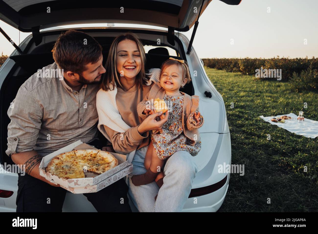 Fröhliche junge Familie, die im Kofferraum des SUV-Autos sitzt und Pizza isst, Mama, Papa und kleine Tochter, die am Wochenende im Freien Spaß haben Stockfoto