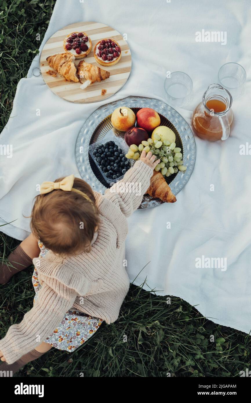 Vertikale flache Lay of Little Baby Girl auf Picknick im Freien, Obst und Kuchen mit Croissants auf dem Plaid bei Sonnenuntergang gelegt Stockfoto