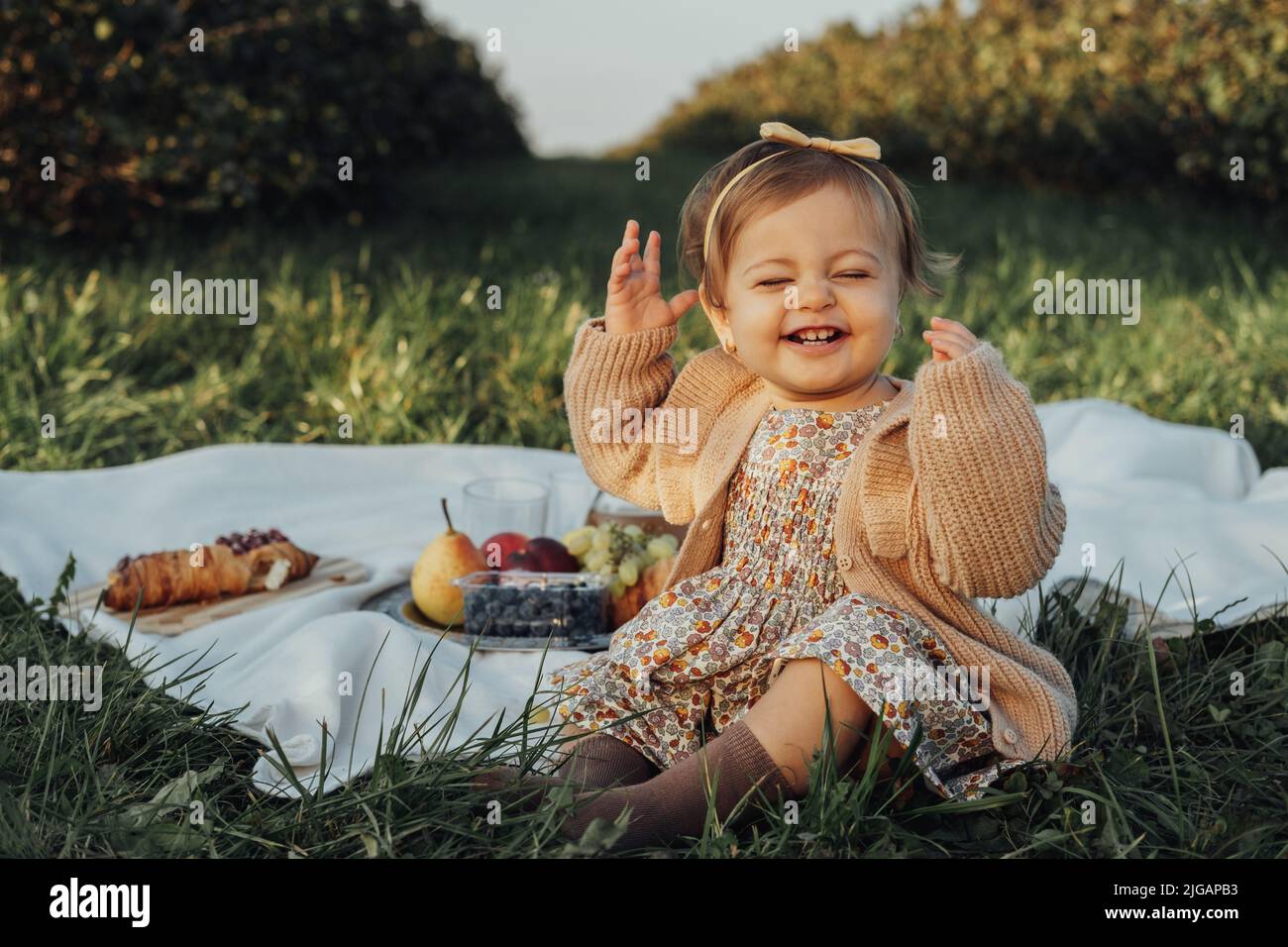 Porträt eines fröhlichen kleinen Mädchens, das auf einem Plaid auf einem Picknick im Freien bei Sonnenuntergang sitzt Stockfoto