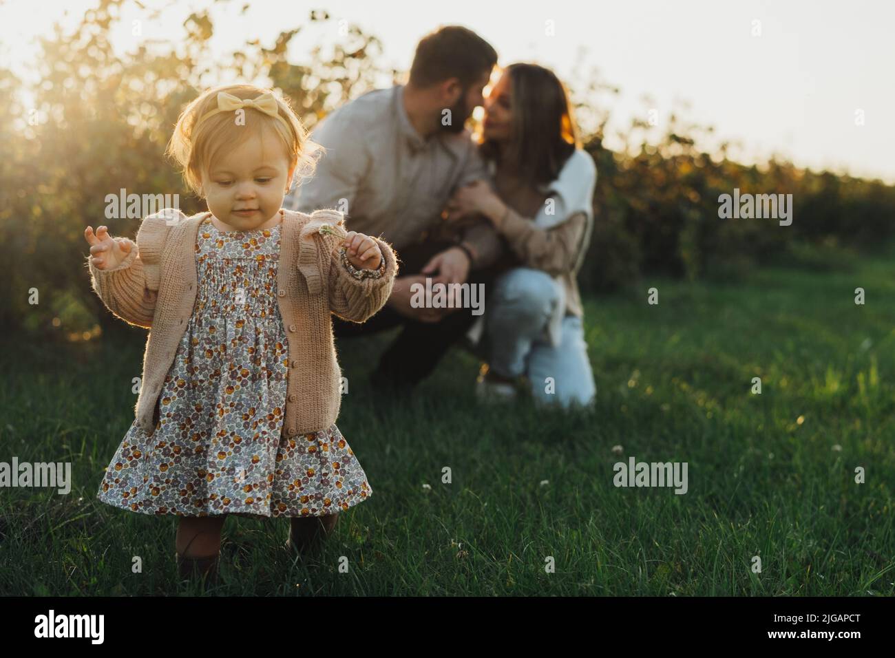 Glückliche junge Familie mit Baby-Tochter im Freien bei Sonnenuntergang, kleines Mädchen macht erste Schritte, während ihre Mutter und Vater auf dem Hintergrund umarmt Stockfoto