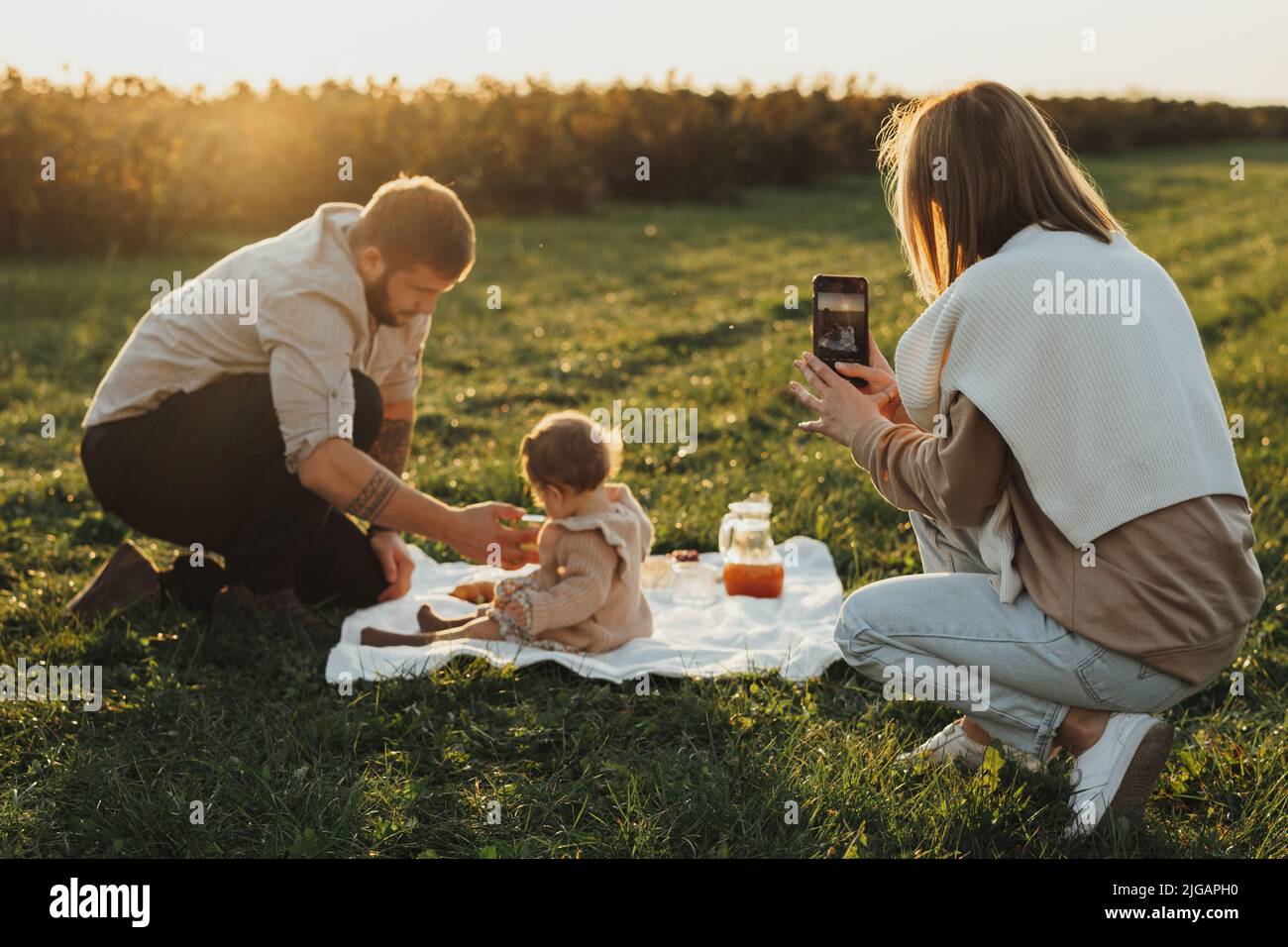 Glückliche junge Familie, die im Freien Picknick macht, Frau, die Fotos von ihrem Mann und ihrer kleinen Tochter bei Sonnenuntergang macht Stockfoto