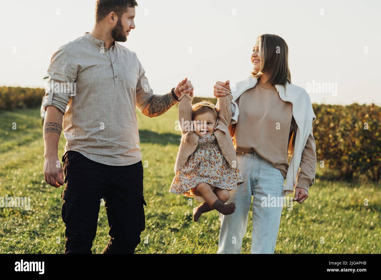 Glückliche junge Familie im Freien, Mutter und Vater heben ihre kleine Tochter bei Sonnenuntergang auf dem Feld mit den Händen Stockfoto
