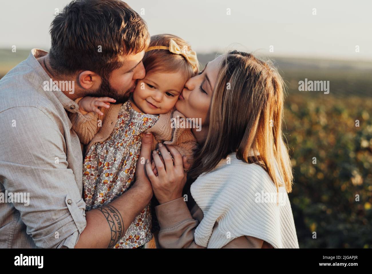 Nahaufnahme Porträt einer glücklichen jungen kaukasischen Familie im Freien, Mutter und Vater küssen ihre kleine Tochter bei Sonnenuntergang Stockfoto
