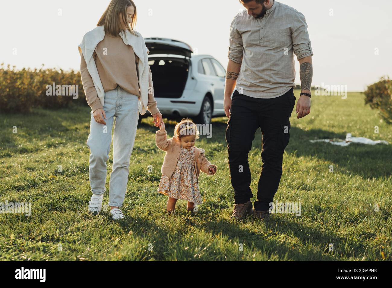 Junge Familie genießt die Fahrt mit dem SUV-Auto, Mutter und Vater mit Baby-Tochter, die bei Sonnenuntergang im Freien auf dem Feld läuft Stockfoto