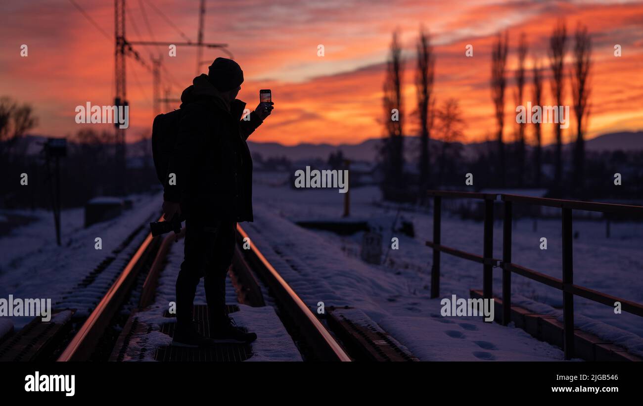 Eine Silhouette eines Mannes auf der Eisenbahnstrecke, der ein Bild eines wunderschönen goldenen Sonnenuntergangs über einer Winterlandschaft fotografiert Stockfoto