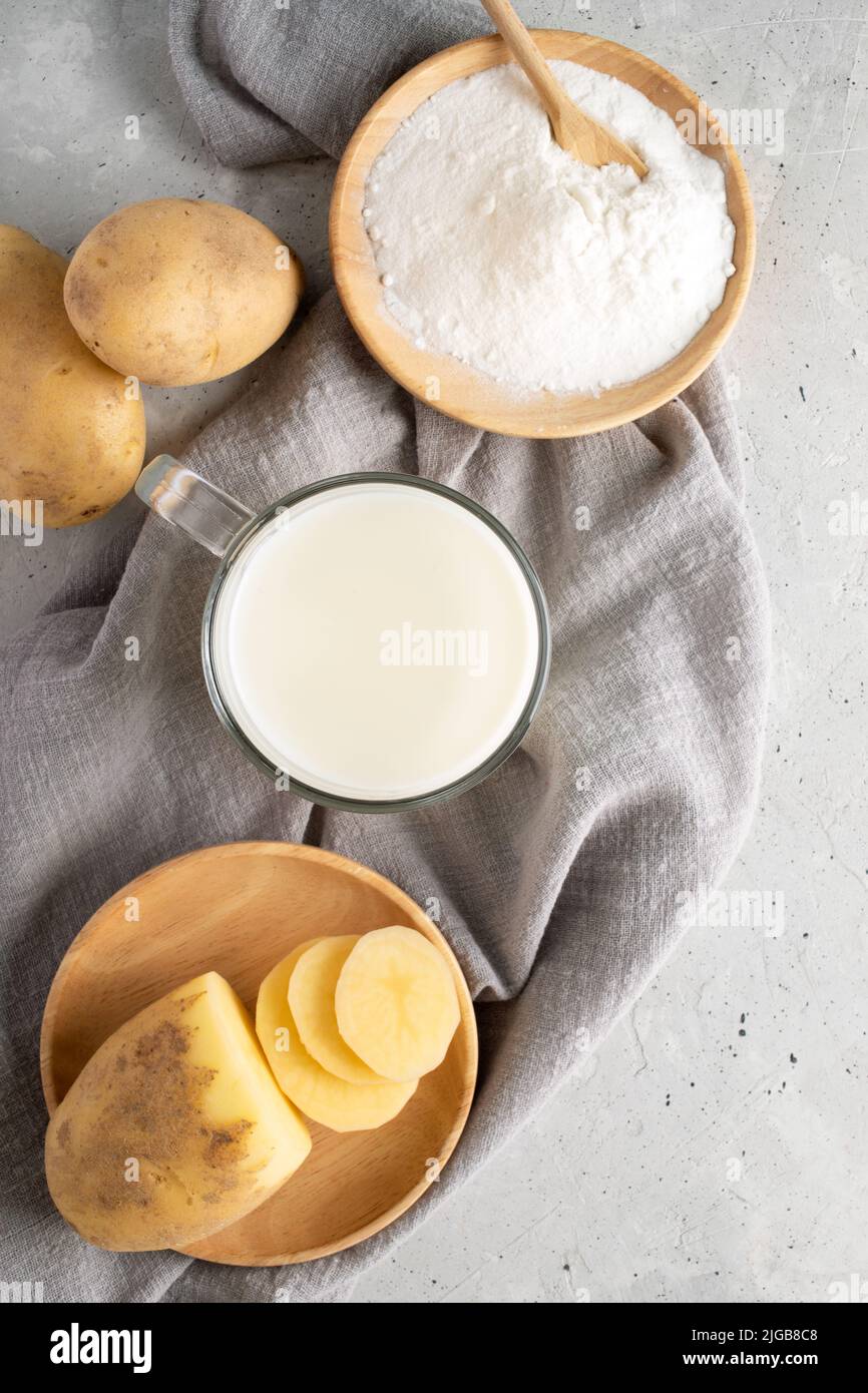 Glasbecher Kartoffelpulver Milch, Holzplatte mit Kartoffelpulver, Knollen auf grauer Serviette auf Beton. Laktosefreie vegane Milch aus Pulver. Anlagenbasis Stockfoto