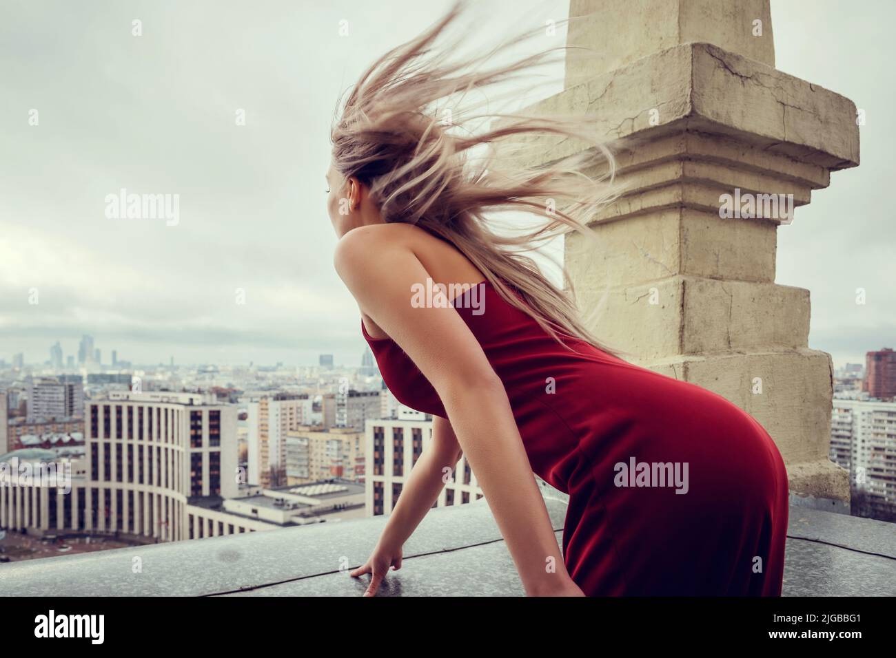 Schlankes Mädchen in rotem Kleid steht auf dem Wolkenkratzer und blickt über die Megacity, die sich bis zum Horizont ausbreitet. Ihr Haar weht im Wind Stockfoto