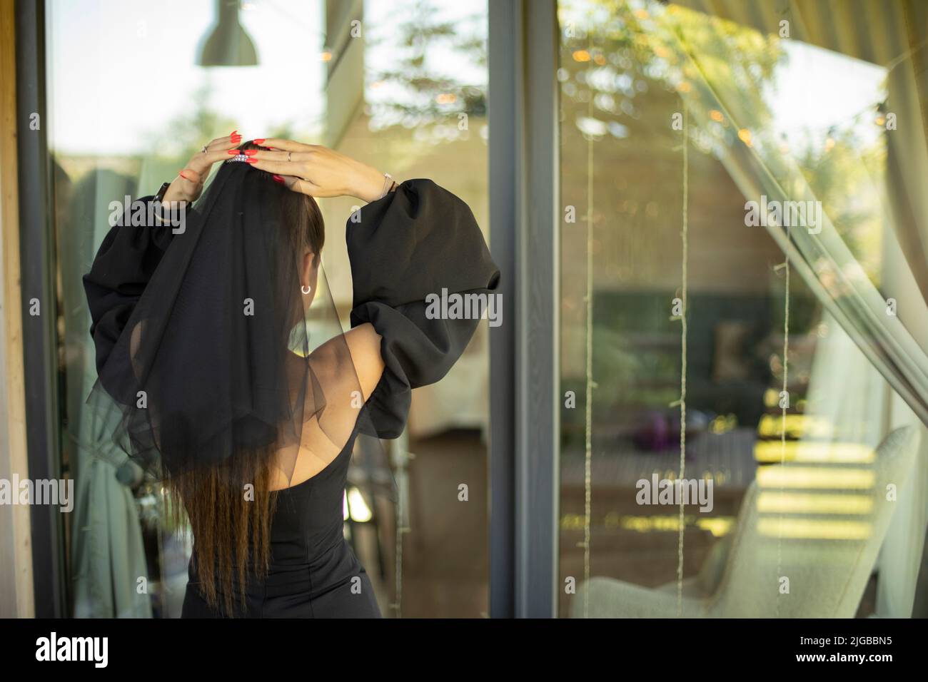 Mädchen in Abendkleid. Schwarzer Schleier. Details der Zeremonie. Mädchen schaut in Spiegelung im Glas. Schwarze Kleidung. Stockfoto