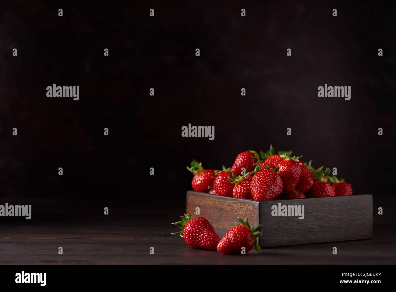 Frische rote reife Erdbeeren in einer quadratischen Holzschüssel auf einem braunen Holztisch. Es gibt zwei Erdbeeren in der Nähe. Brauner Hintergrund, horizontal. Nahaufnahme Stockfoto