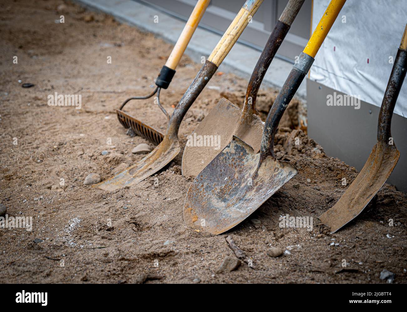 Schmutzige gebrauchte Schaufeln und ein Rechen lehnen sich während einer Arbeitspause auf einer neuen Baustelle an eine Wand. Stockfoto