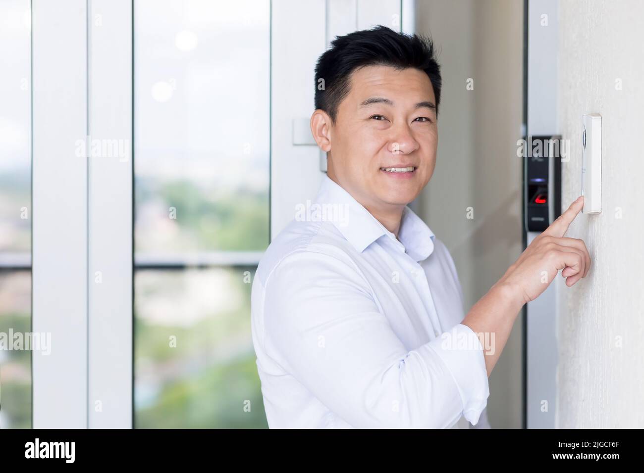 Porträt des asiatischen Geschäftsmannes, der Mann tritt in ein modernes Büro, verwendet eine Türklingel mit Fingerabdruckschloss, der Mann blickt auf die Kamera und lächelt Stockfoto