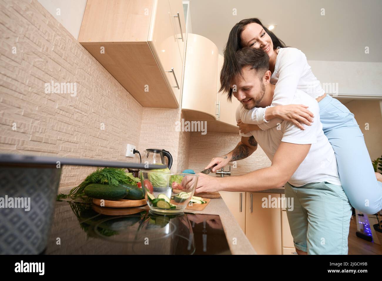 Ehemann hob seine Frau auf dem Rücken, während er Salat kochte Stockfoto