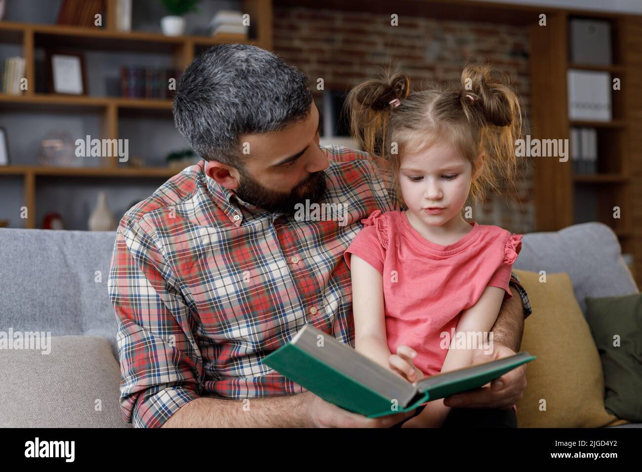 Zu Hause lasen der junge Vater und die junge Tochter Interessantes Buch. Liebende Eltern bärtiger Mann lehrt sein Vorschulkind zu lesen. Dvory liest Märchen Geschichte Stockfoto