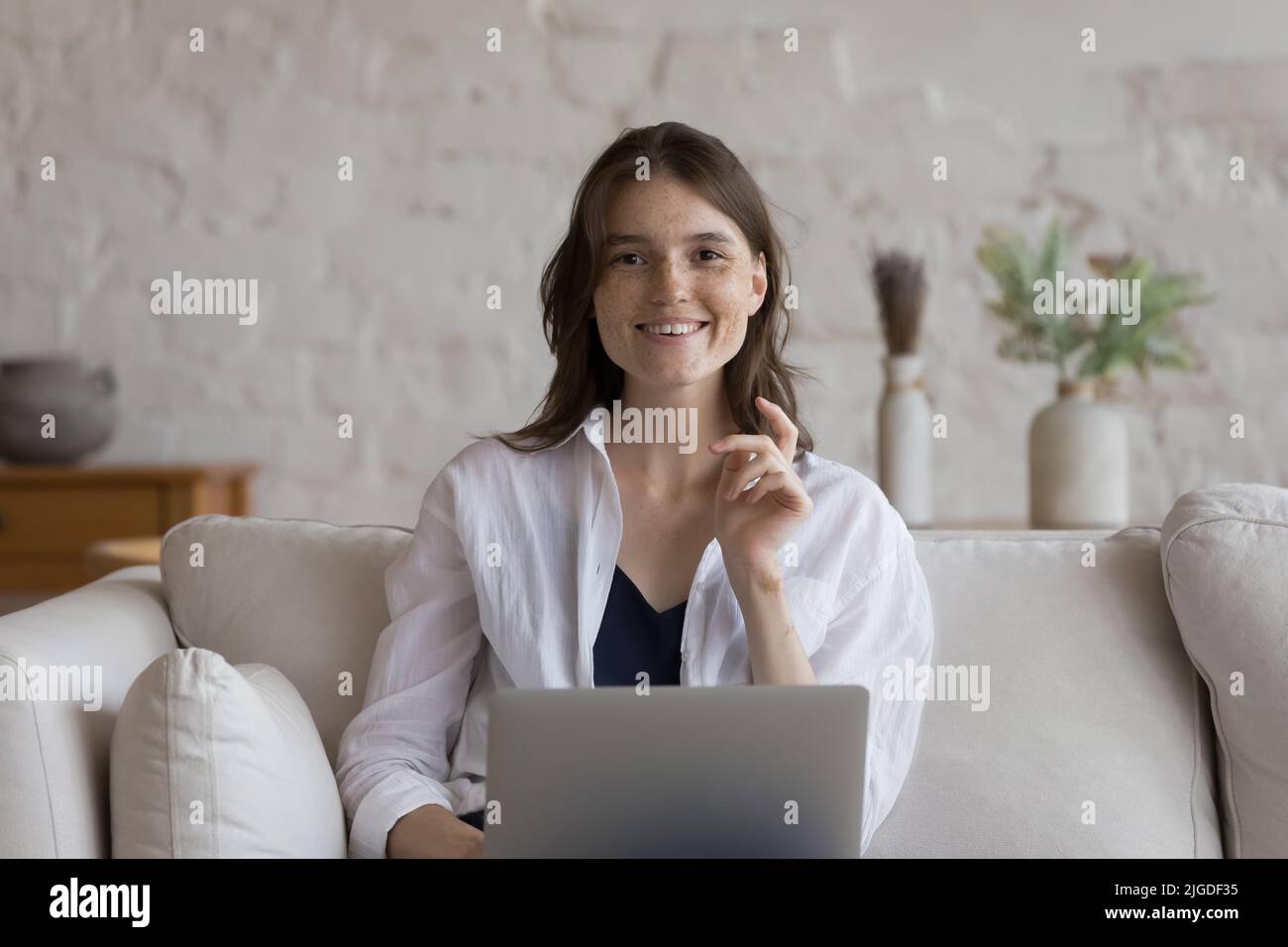 Die junge Frau sitzt auf dem Sofa, während der Laptop die Kamera anschaut Stockfoto