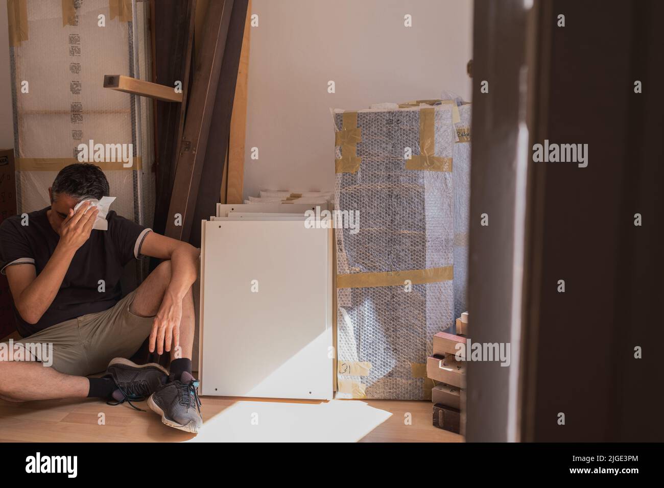 Umzug in ein neues Zuhause. Der müde Mann, der auf dem Boden sitzt, wischt seinen Schweiß mit einem Papierhanky ab. Pause nach dem Transport von verpacktem Material und Möbeln Stockfoto