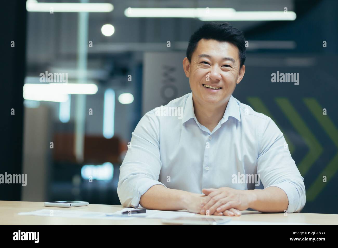 Asiatischer Geschäftsmann im modernen Büro mit Blick auf die Webcam, lächelnd am Tisch sitzend, Online-Meeting Stockfoto