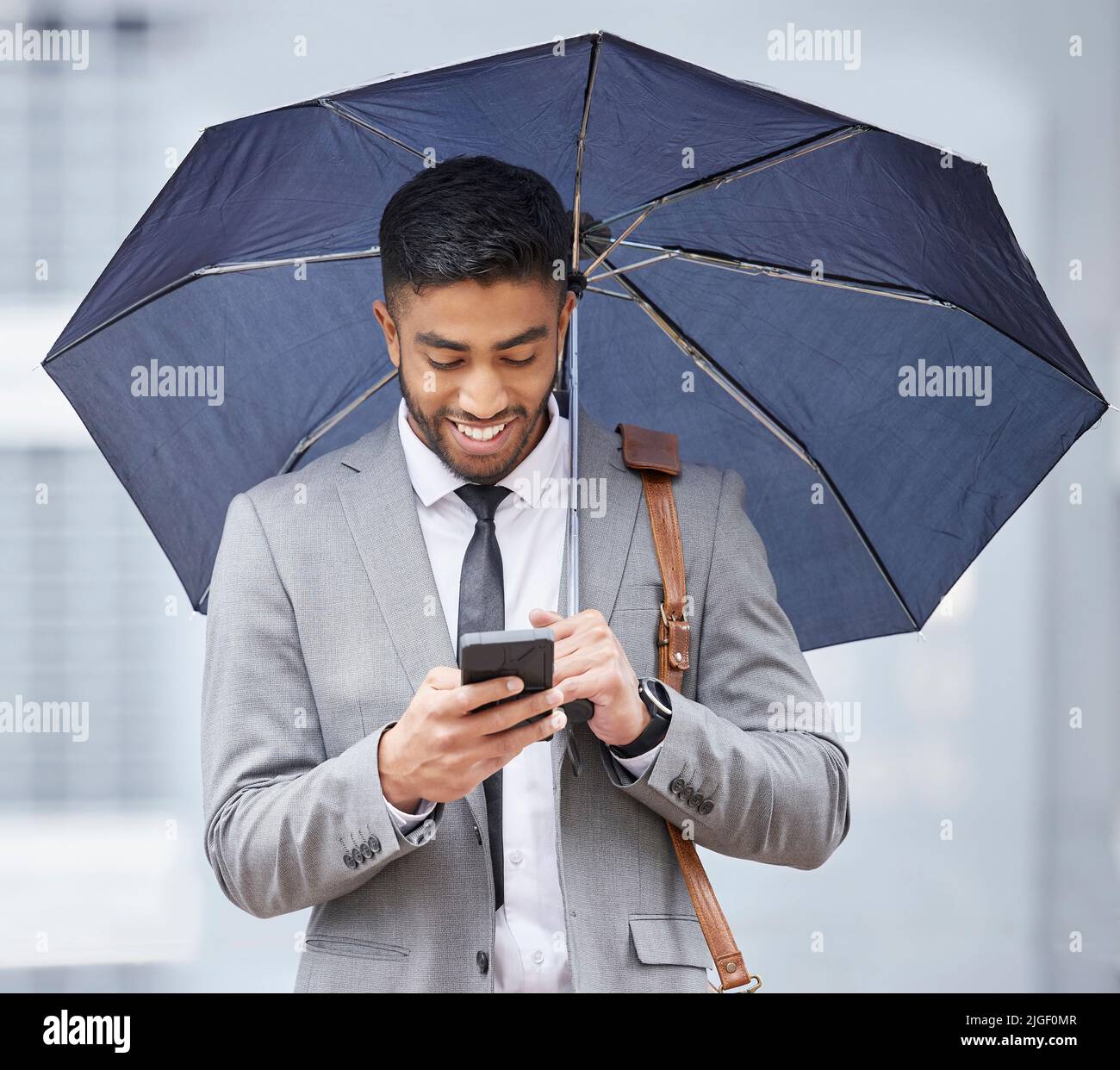 Er hat Geschäfte aus allen Blickwinkeln. Ein junger Geschäftsmann hält einen Regenschirm, während er an einem regnerischen Tag in der Stadt ein Handy benutzt. Stockfoto