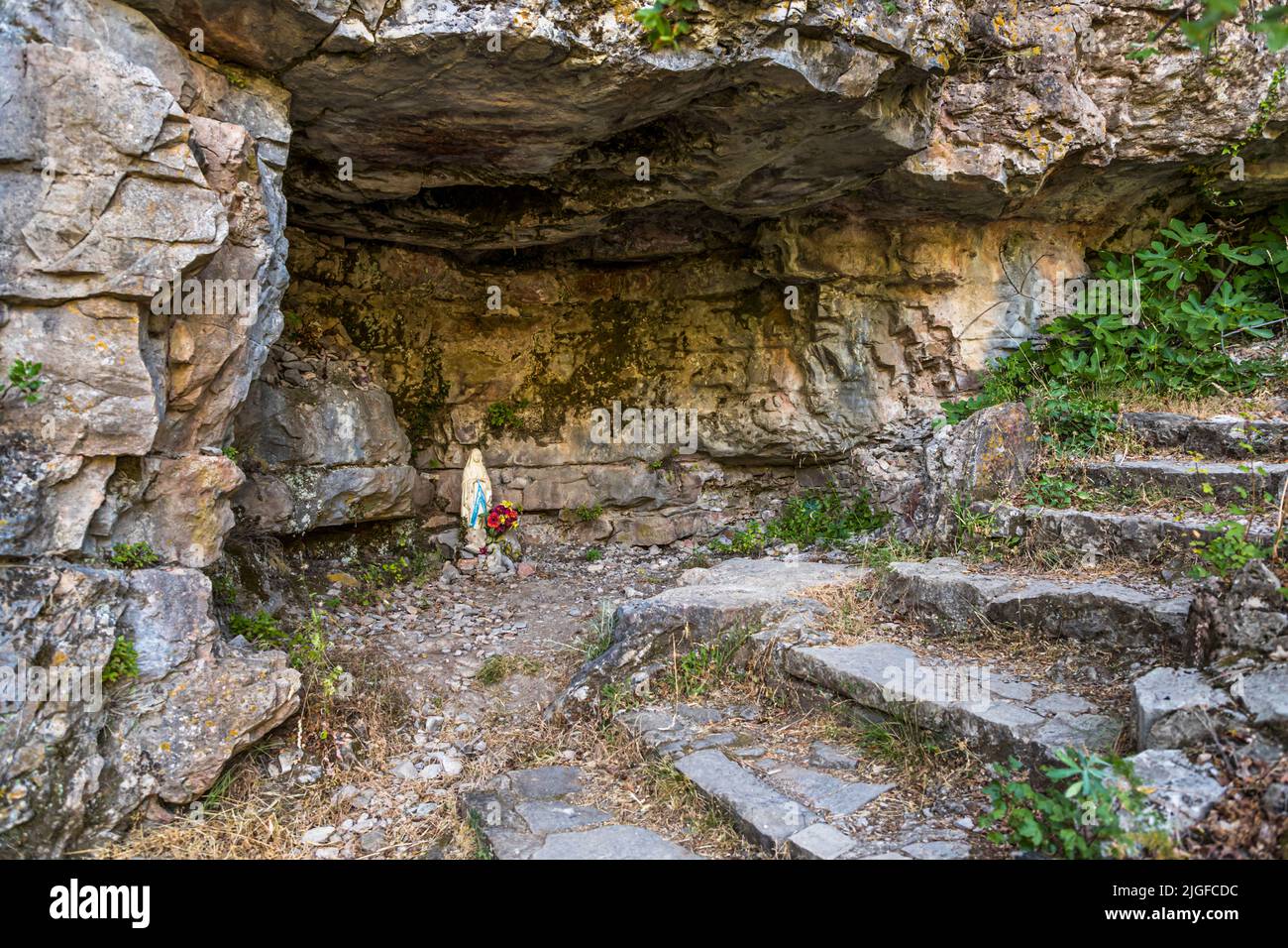 Eine Statue der Madonna, in Tuch gehüllt und mit Blumen geschmückt, steht in einer Felsspalte entlang des Wanderwegs im Cirque de Navacelles nahe Lodève, Hérault, Frankreich. Die überhängende Kalksteinwand bildet eine natürliche Grotte über dem kleinen Wegeheiligtum. Stockfoto