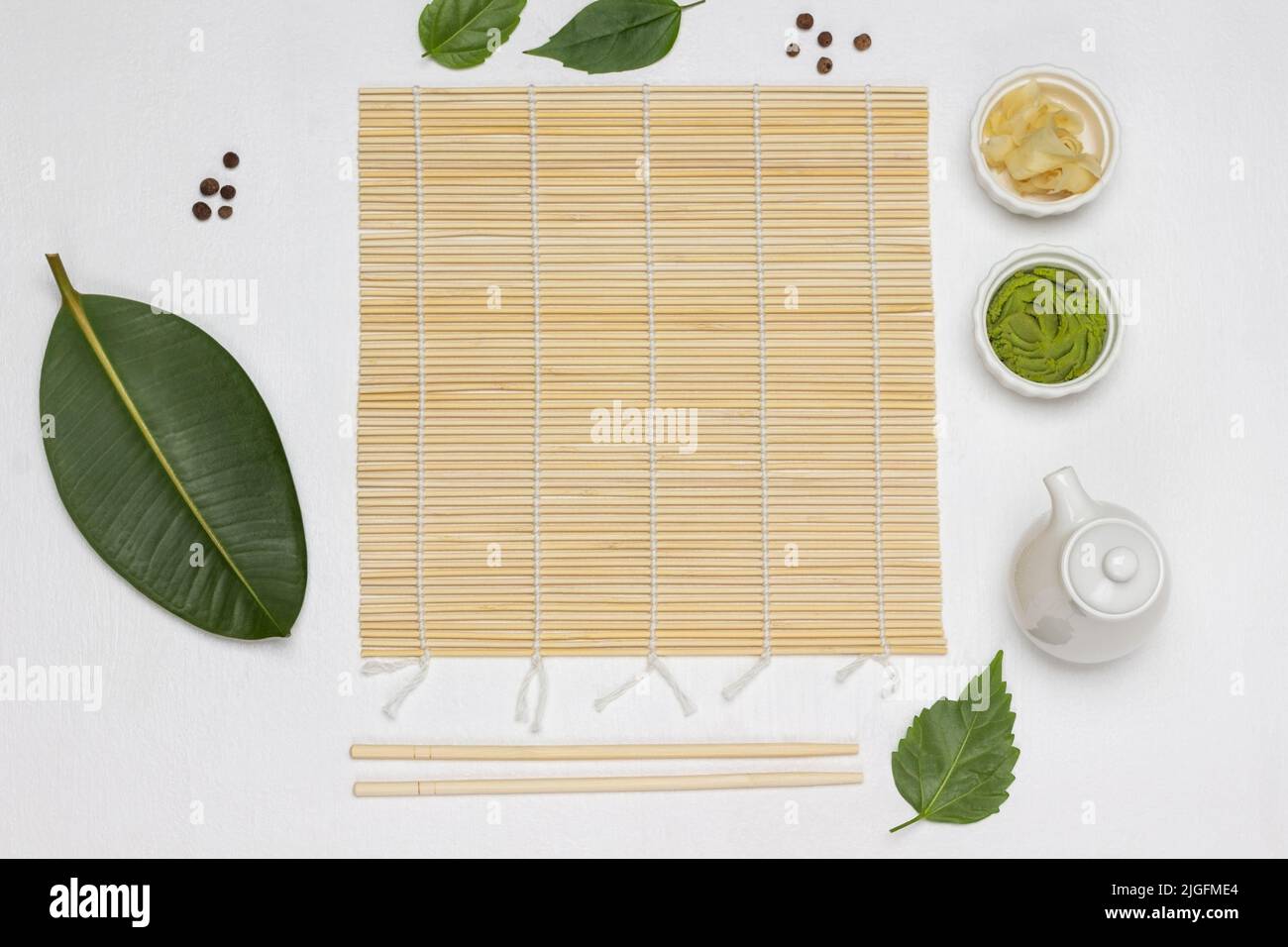 Bambusmatte, Essstäbchen, Sushi-Gewürze und grüne Blätter auf dem Tisch. Flach liegend. Weißer Hintergrund Stockfoto