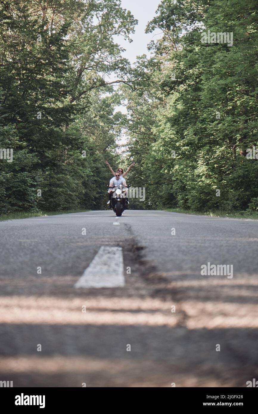 Ein junges glückliches Paar fährt mit dem Motorrad auf einer asphaltierten Straße im Wald, Freiheit und Geschwindigkeit Stockfoto