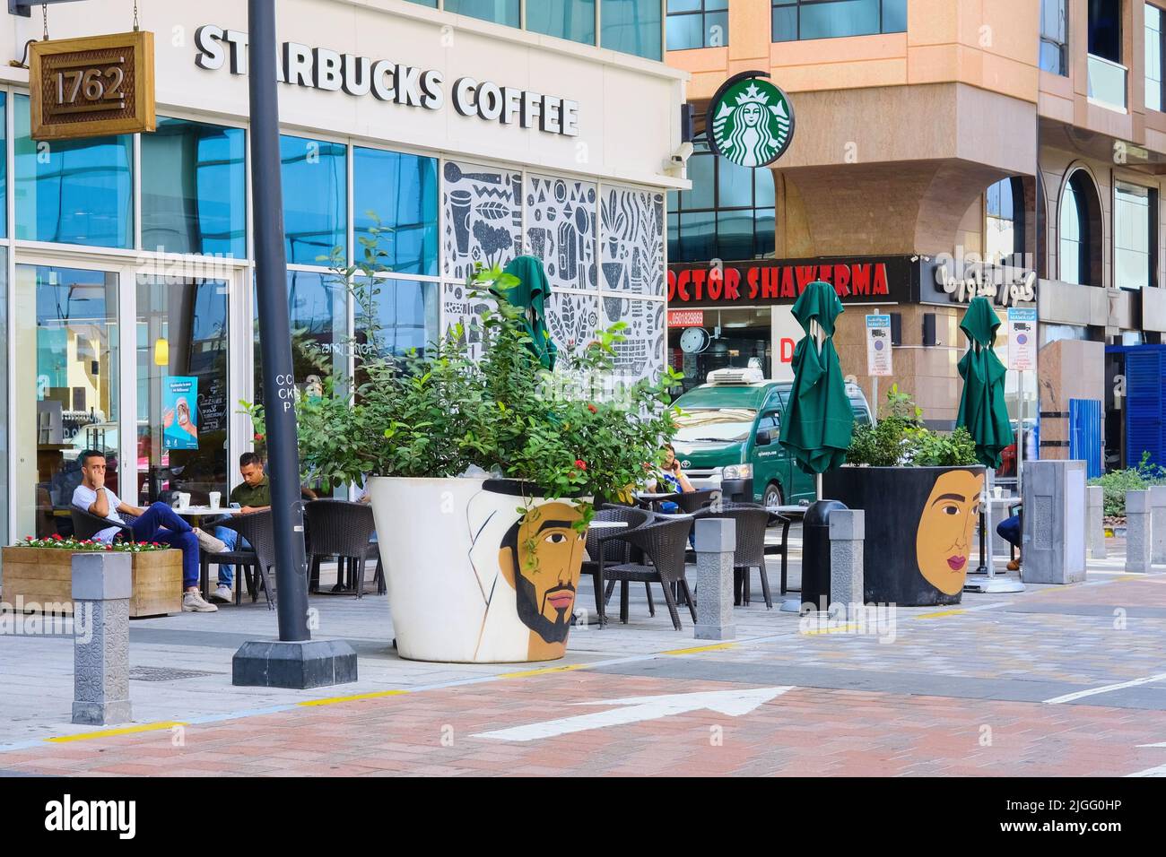 Blick auf die Straße mit Terrasse des Starbucks Cafés mit riesigen Blumenvasen mit menschlichen Gesichtern, arabischer Frauenmann, im Vordergrund. Stockfoto
