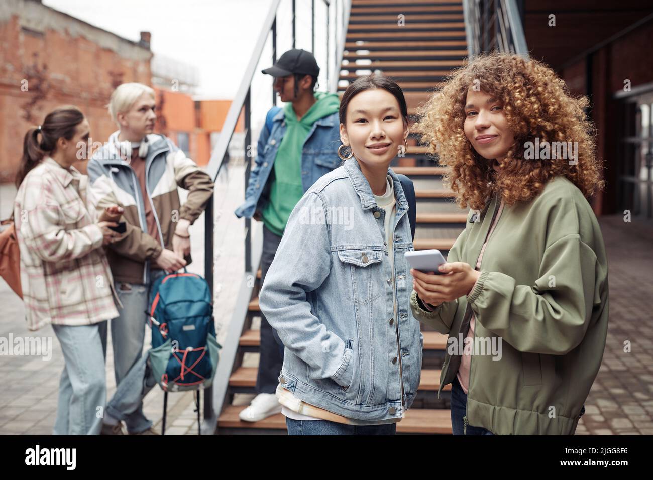 Zwei Jugendliche interracial Mädchen in Casualwear stehen vor ihren Freunden, die nach dem Unterricht in der Hochschule gegen die Treppe plaudern Stockfoto