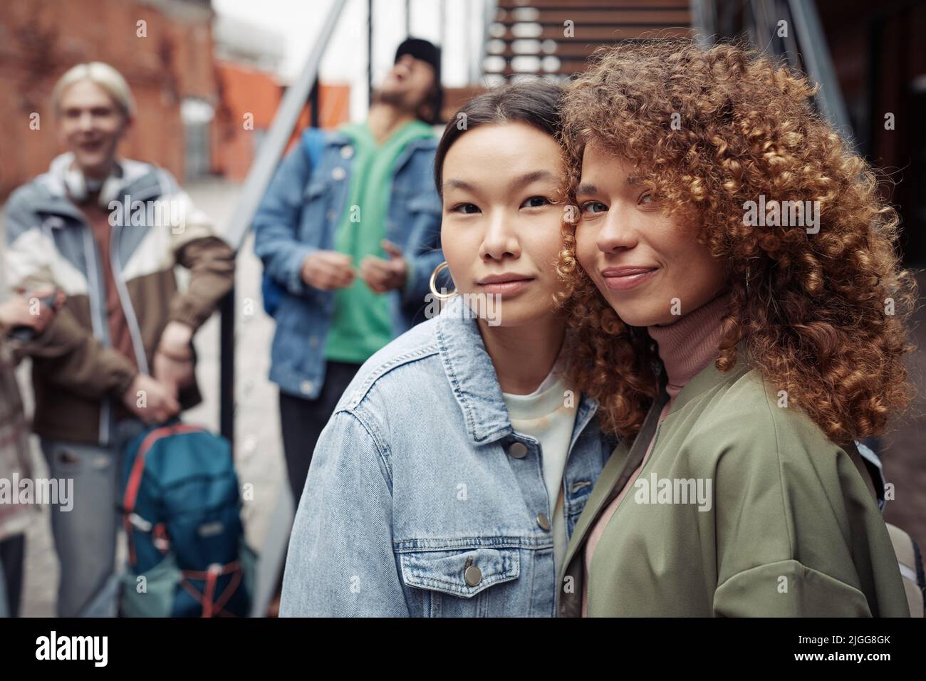 Zwei fröhliche junge Frauen in stilvoller Casualwear blicken auf die Kamera, während sie sich im Freien mit Teenagern unterhalten, die sich über die Treppe unterhalten Stockfoto