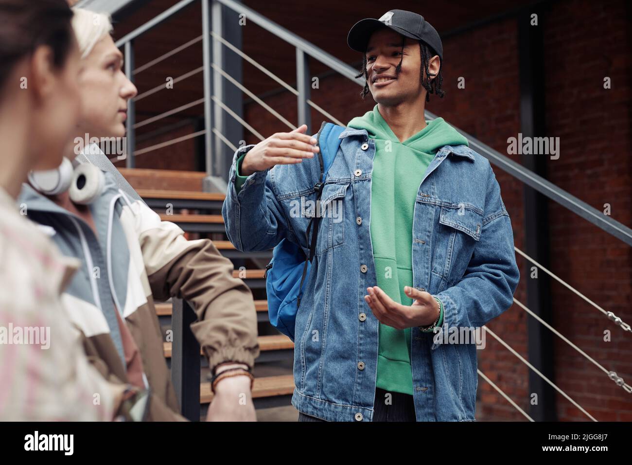 Junger lächelnder schwarzer Mann in Casualwear, der auf der Treppe an der Backsteinmauer des Gebäudes steht und sich nach dem Unterricht mit seinen Kollegen unterhält Stockfoto