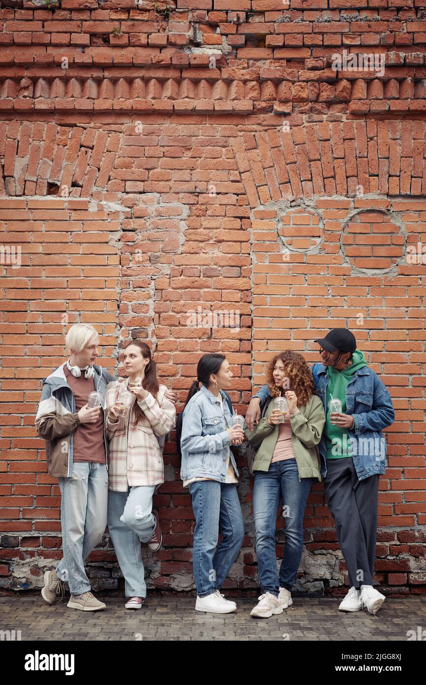 Eine Gruppe von multikulturellen Teenagern und Mädchen in Casualwear, die gegen eine Ziegelsteinmauer stehen, sich nach dem College mit Soda unterhalten und sich unterhalten Stockfoto