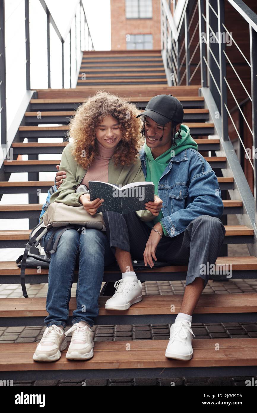 Lächelndes Teenager-Paar in Casualwear, das in einem Copybook die Vorlesungsnotizen durchschaut, während es auf einer langen Treppe am modernen Gebäude sitzt Stockfoto