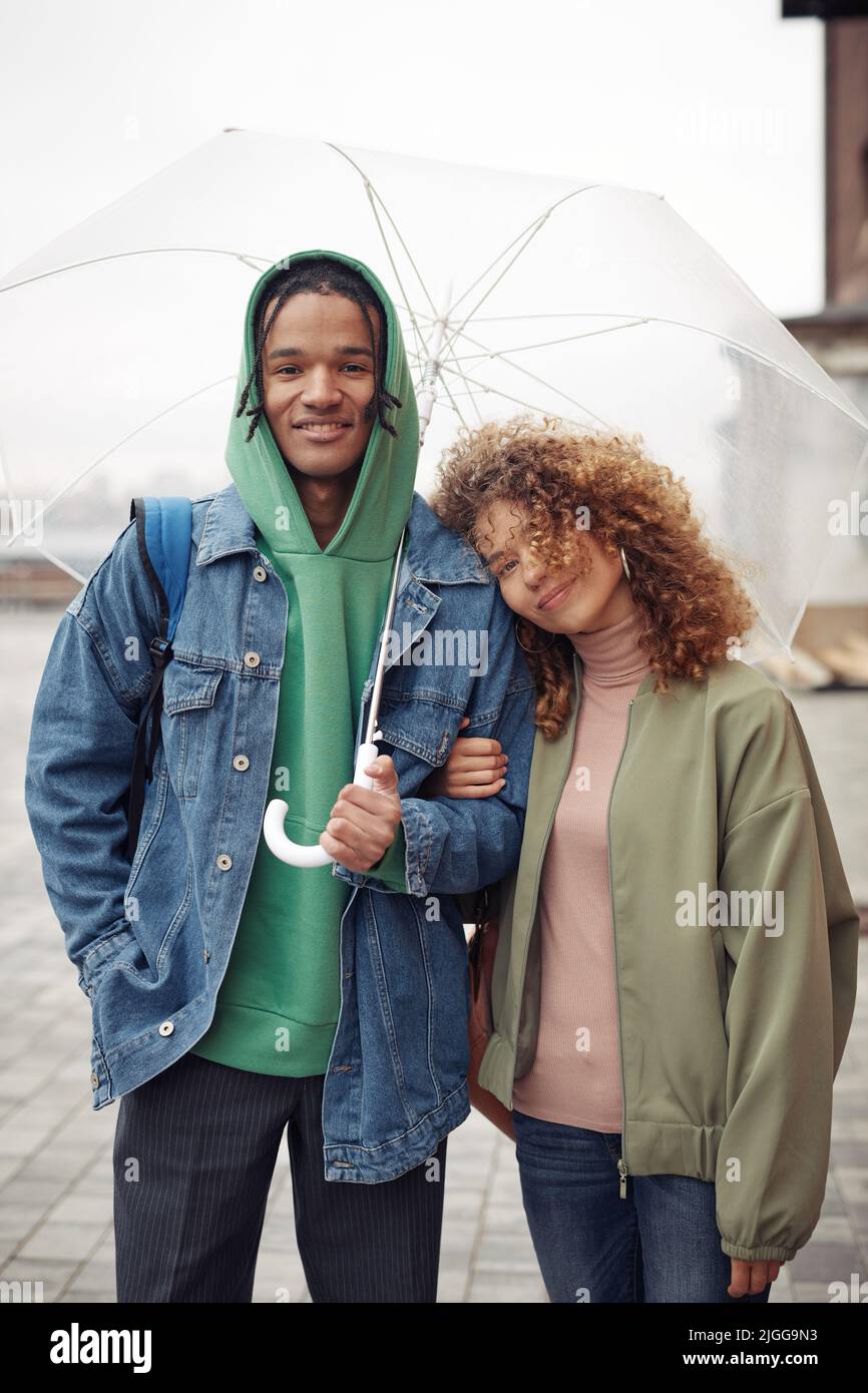 Glückliches junges interrassisches Paar in Casualwear, das unter einem Regenschirm dicht beieinander steht und beim Gehen auf die Kamera schaut Stockfoto