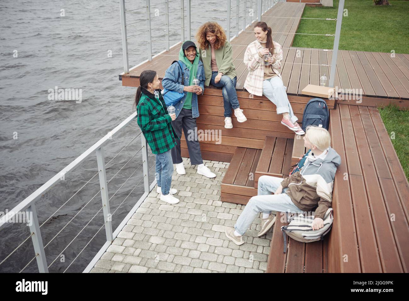 Eine Gruppe von multikulturellen Teenagern in Casualwear, die sich am Flussufer unterhalten und entspannen, während einige von ihnen auf einer Holzbank sitzen Stockfoto