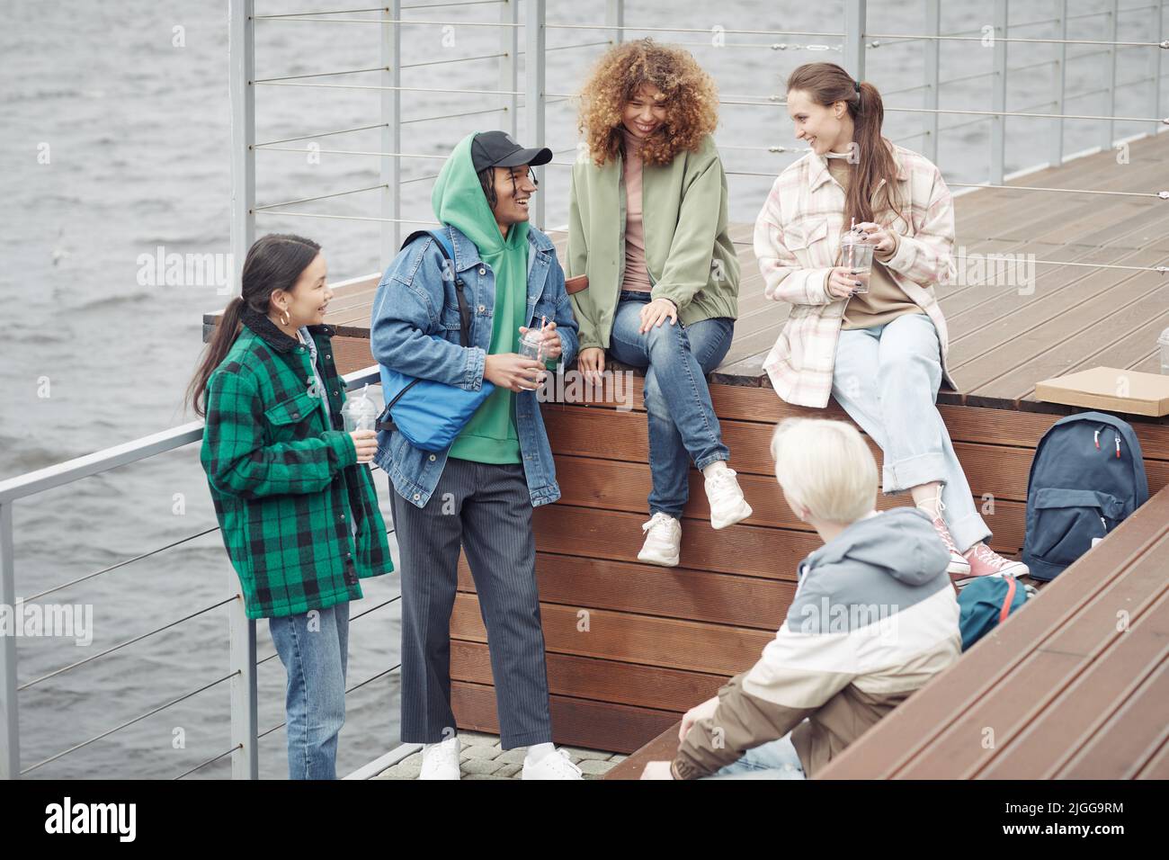 Fünf fröhliche Teenager-Freunde in Casualwear erzählen Witze oder diskutieren lustige Dinge, während sie sich nach dem College auf dem hölzernen Pier am Wasser ausruhen Stockfoto