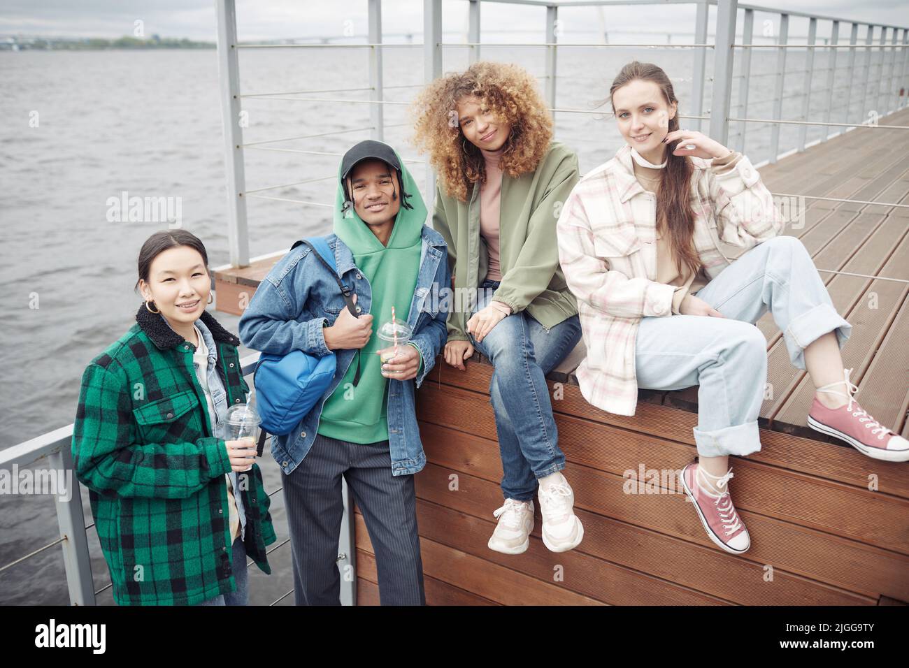 Vier fröhliche und freundliche Teenager in legerer Kleidung, die am Wochenende oder nach dem Unterricht in der Schule auf dem hölzernen Pier die Kamera betrachten Stockfoto