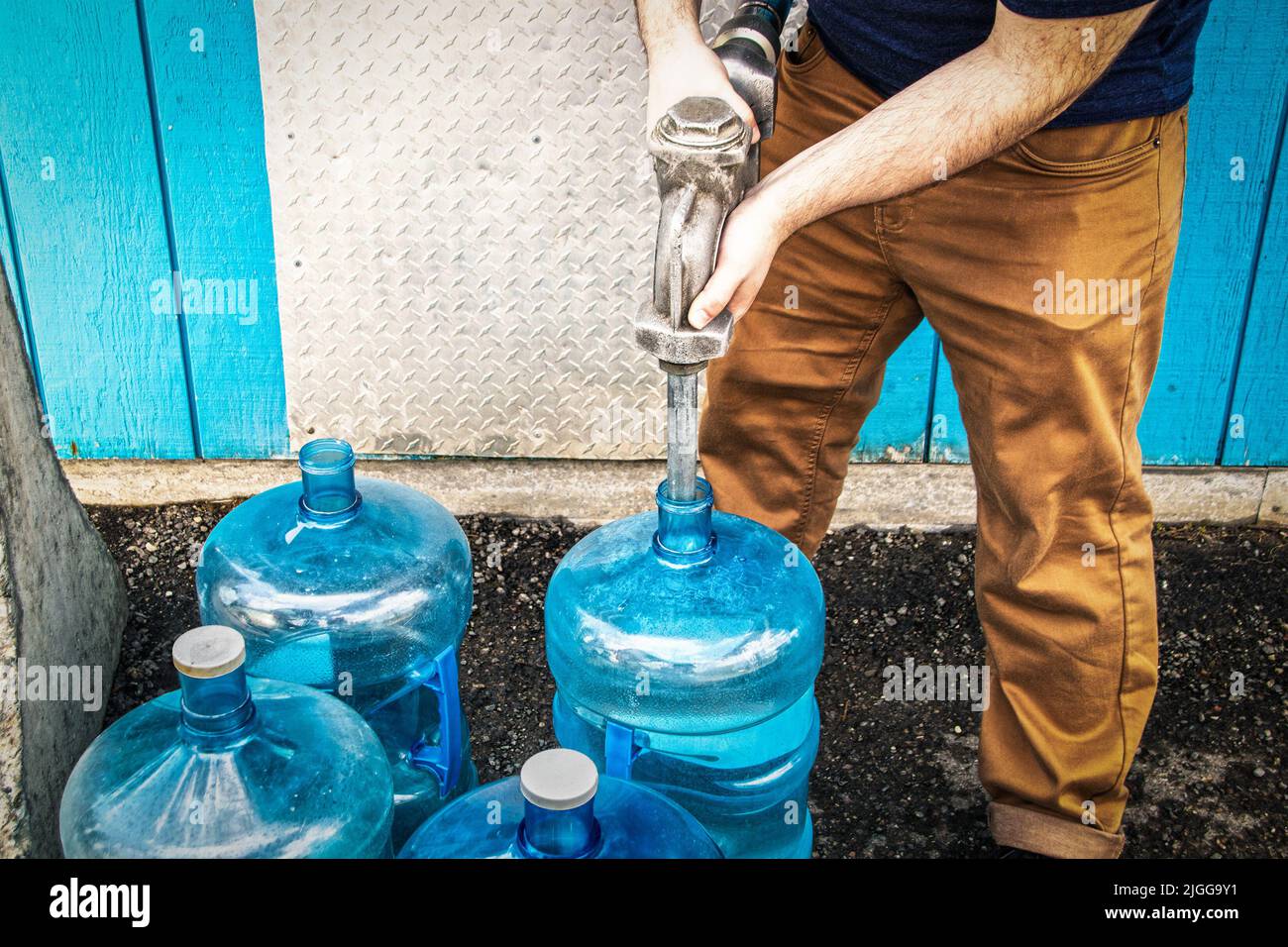Mann in Arbeitshose mit Metalldüse, der blaue Flaschen mit Wasser füllt, um sie in der Kabine ohne fließendes Wasser zu verwenden - neben dem Gebäude auf rauer Rückseite stehend Stockfoto