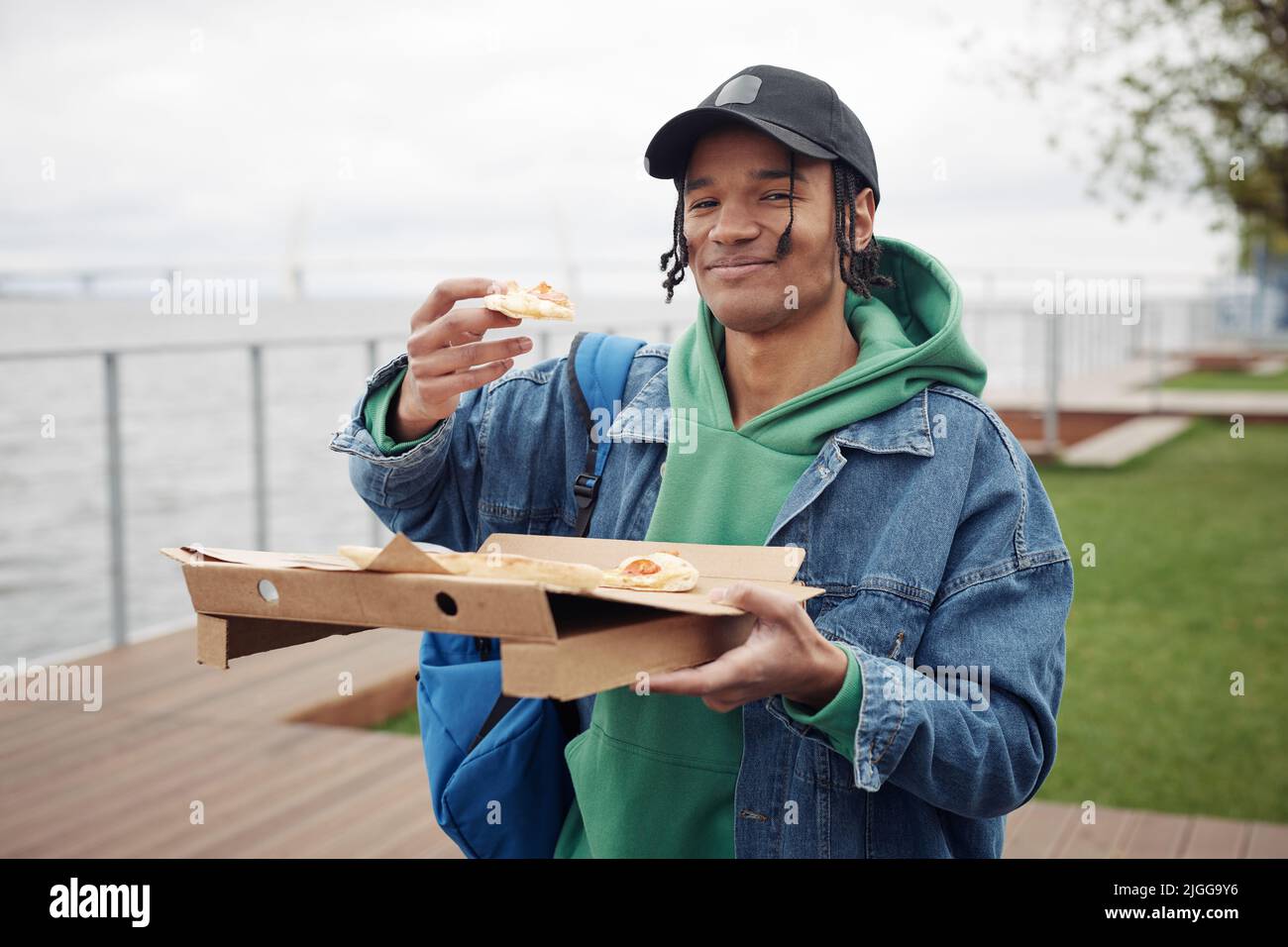 Fröhlicher schwarzer Kerl in der Freizeitkleidung, der ein Stück Pizza über einer quadratischen Schachtel hält, während er einen Snack hat und während des Picknicks im Park die Kamera anschaut Stockfoto