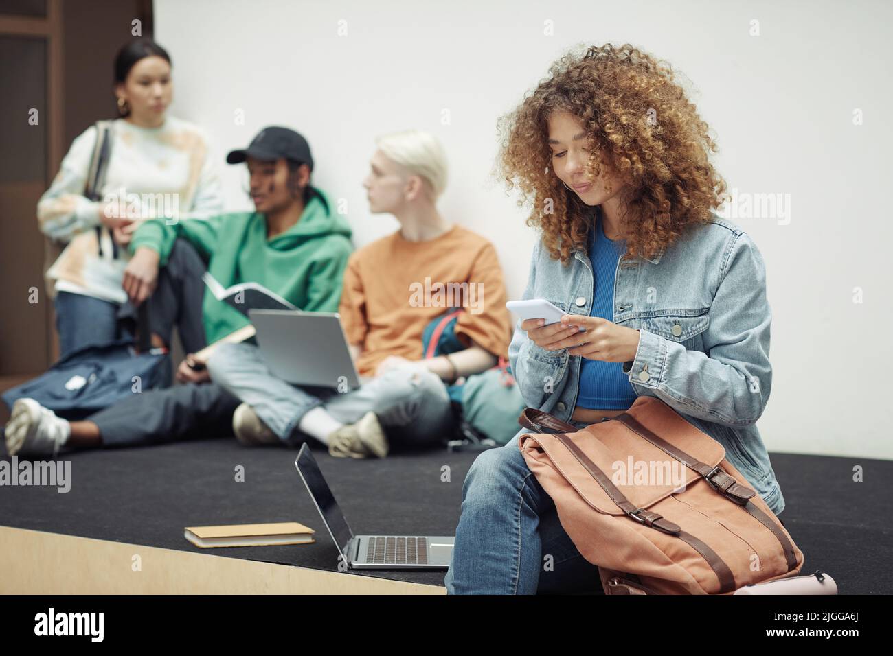 Hübsches Teenager-Mädchen in Casualwear, das in der Pause in einem Smartphone schreibt, während sie im Hörsaal sitzt, während ihre Gruppenmitglieder über Vortragsnotizen diskutieren Stockfoto