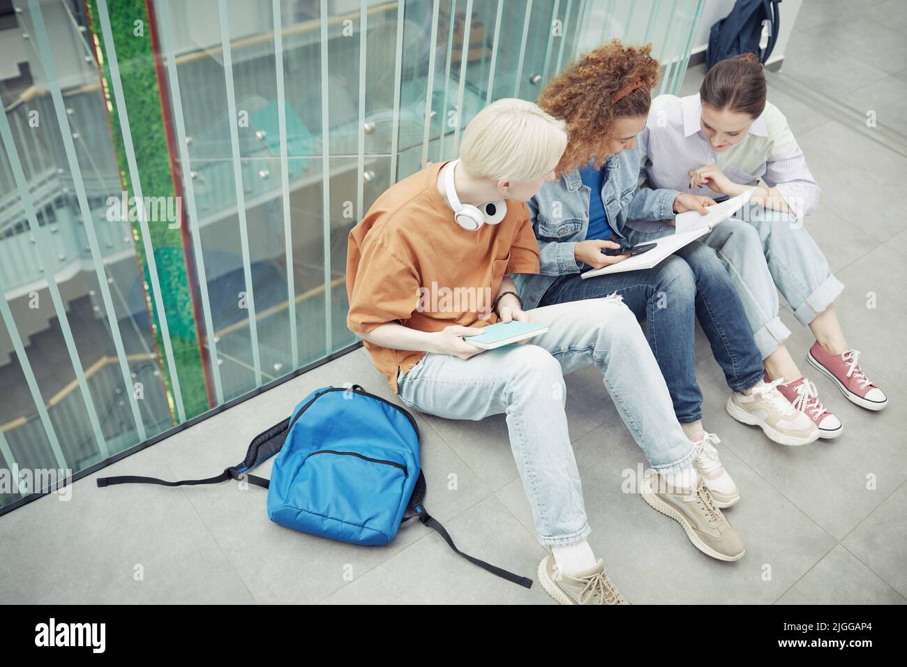 Drei interkulturelle Jugendliche in Casualwear schauen sich im Copybook eines Mädchens mit welligen Haaren Notizen an, während sie sich auf dem Boden ausruhen Stockfoto