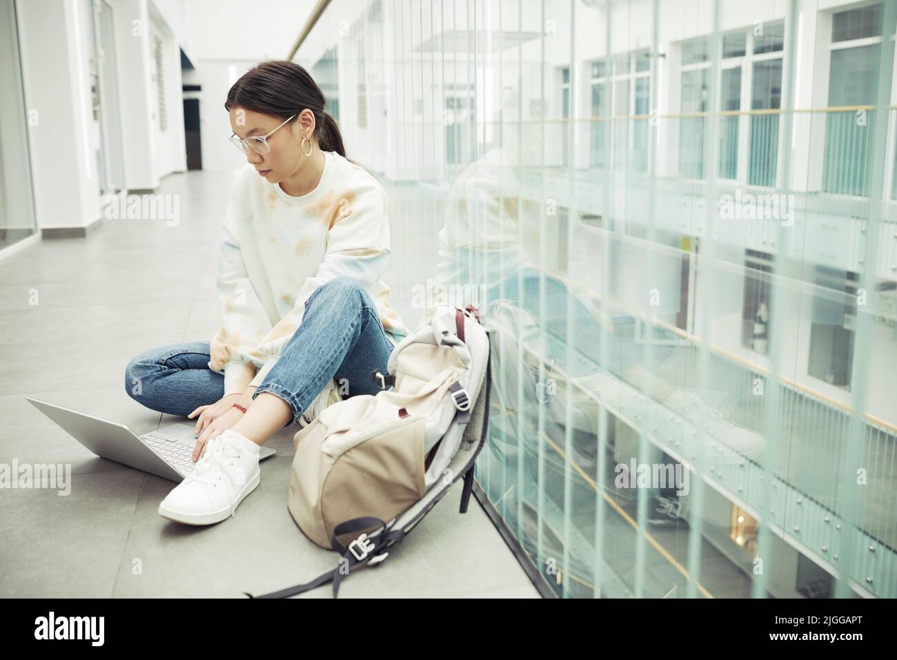 Serious Teenager Mädchen asiatischer Ethnie in Casualwear sitzen auf dem Boden der College-Korridor und suchen durch Online-Informationen Stockfoto