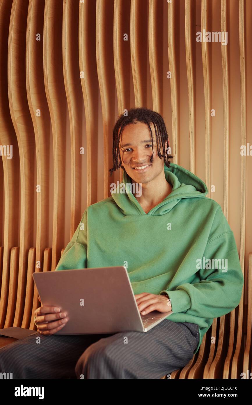 Glücklicher afroamerikanischer Teenager-Typ im grünen Hoodie, der auf dem Laptop tippt und die Kamera anschaut, während er an der Wand im College-Korridor sitzt Stockfoto