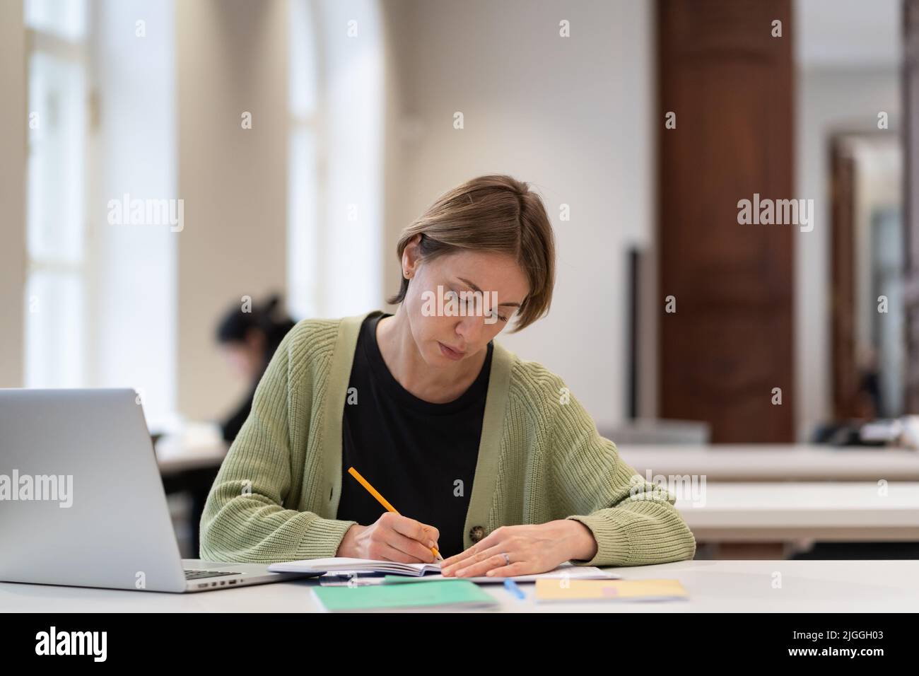 Studentin mittleren Alters, die sich auf die Prüfung in der öffentlichen Bibliothek vorbereitet, während sie mit einem Laptop am Tisch sitzt Stockfoto