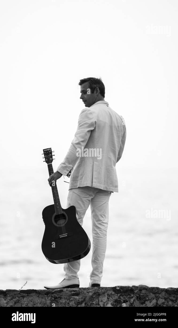 Galle, Sri Lanka - 02 15 2022: Weiß gekleideter Mann, der eine Gitarre in der Hand hält und auf den Strand zurückblickt Schwarz-Weiß-Foto. Stockfoto