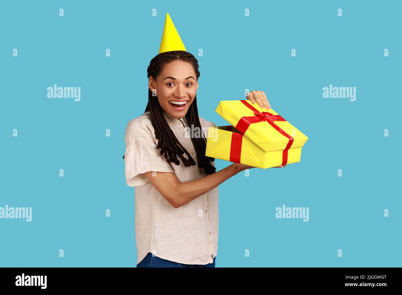 Porträt einer glücklichen, staunenden Frau in gelbem Partykegel, die in die Geschenkbox schaut, das Geschenk öffnet und mit Glück die Kamera anschaut, das weiße Hemd trägt. Innenaufnahme des Studios isoliert auf blauem Hintergrund. Stockfoto