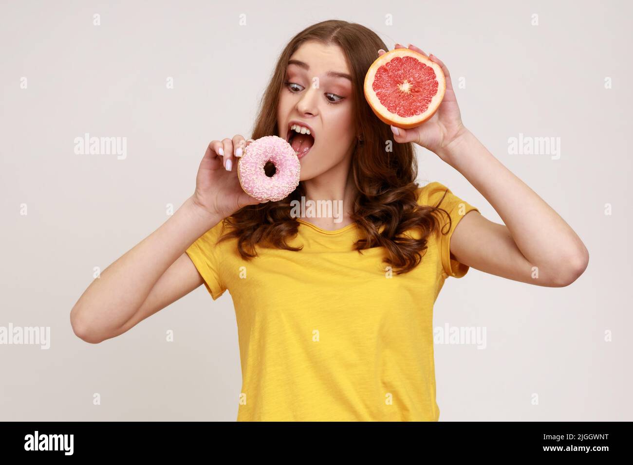 Hungriges Teenager-Mädchen in gelbem T-Shirt beißt süßen Junk-Donut anstelle von frischen saftigen Orange, die Wahl Junk-Food, Mangel an Willenskraft und Motivation. Innenaufnahme des Studios isoliert auf grauem Hintergrund. Stockfoto