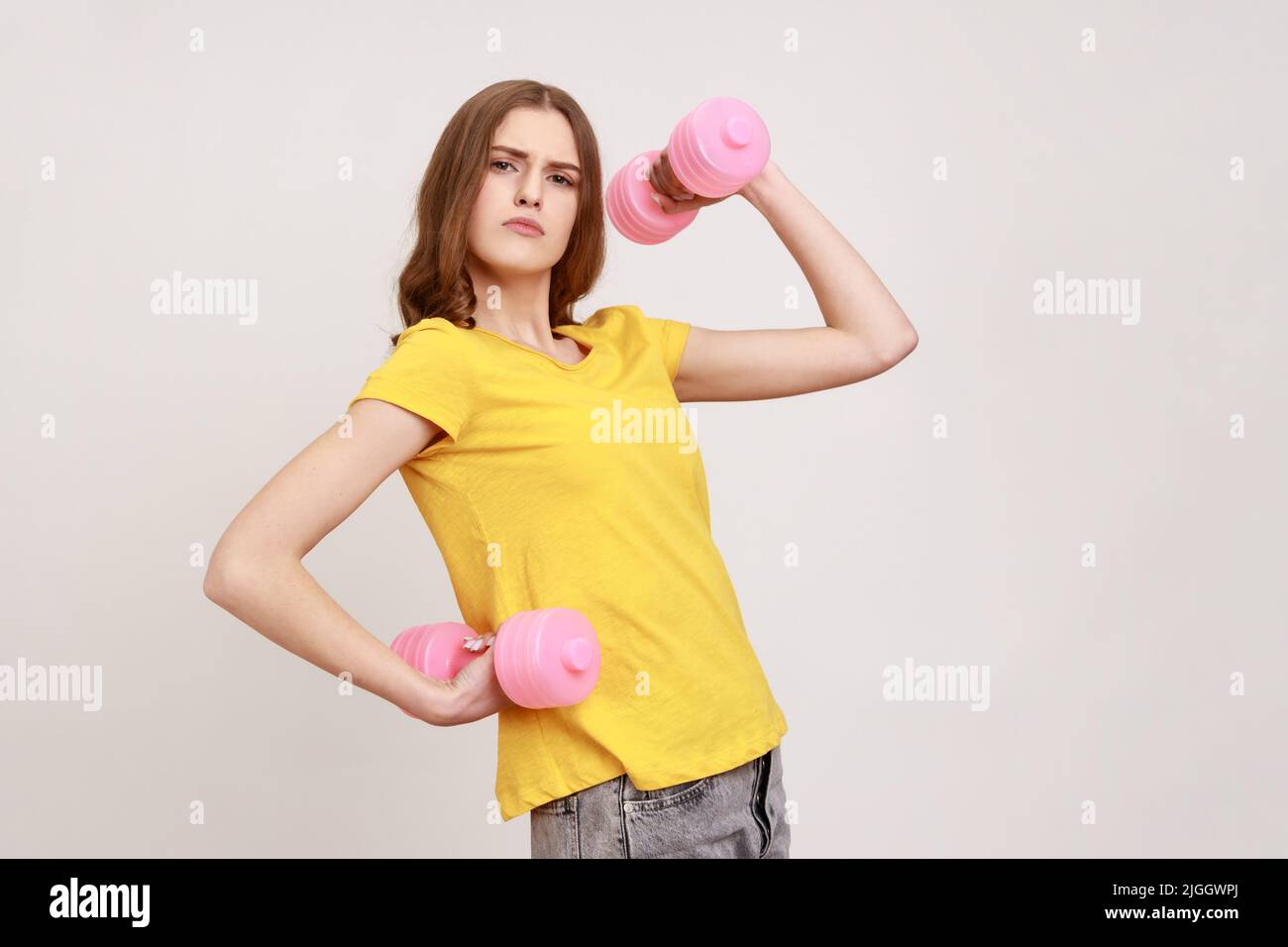 Durchsetzungsfähig lustige Teenager-Mädchen mit braunen Haaren in gelben casual T-Shirt aufsteigenden Händen halten kleine rosa Hanteln, Pumpen bis Muskeln. Innenaufnahme des Studios isoliert auf grauem Hintergrund. Stockfoto