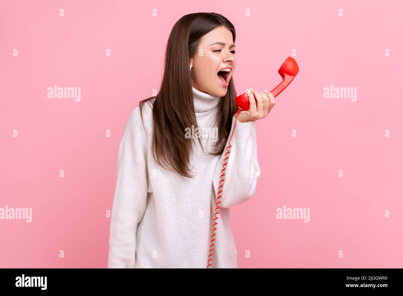 Wütend junge Brünette Frau schreiend auf roten Retro-Hörer, zum Ausdruck Aggression, ärgerlich Client, trägt weißen lässigen Stil Pullover. Innenaufnahme des Studios isoliert auf rosa Hintergrund. Stockfoto