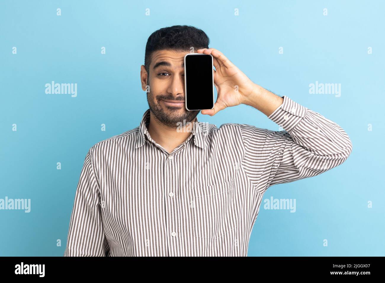 Porträt eines lächelnden, gutaussehenden Geschäftsmannes, der mit dem Smartphone ein Auge hält und bedeckt, die Kamera anschaut und ein gestreiftes Hemd trägt. Innenaufnahme des Studios isoliert auf blauem Hintergrund. Stockfoto